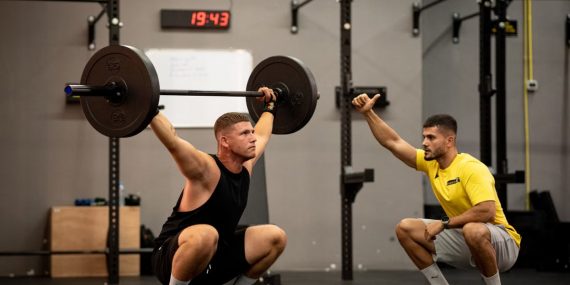 Athlete performing weightlifting exercise under trainer's guidance in a gym setting.